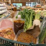 岐阜県のローカルスーパーで、買い物かごに明宝ハム風のハム、鶏ちゃん風の味付け肉、五平餅のたれ、朴葉みそ風の商品、地元野菜が入っている自然な構図
