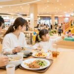 ショッピングモールのフードコートで、家族がランチを楽しみながら子どもが近くのキッズスペースを眺めている